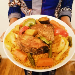 une serveuse présente un plat de thiéboudienne sénégalais, dit encore tiep ou tchep. Le poisson et les légumes sont posés sur le riz à base de sauce tomate.